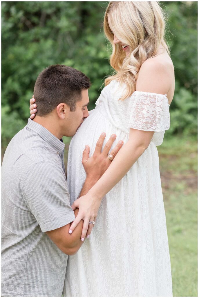 Pregnant mom wears a beautiful, off the shoulder, white dress and the soon to be Dad, bends down and kisses her belly.