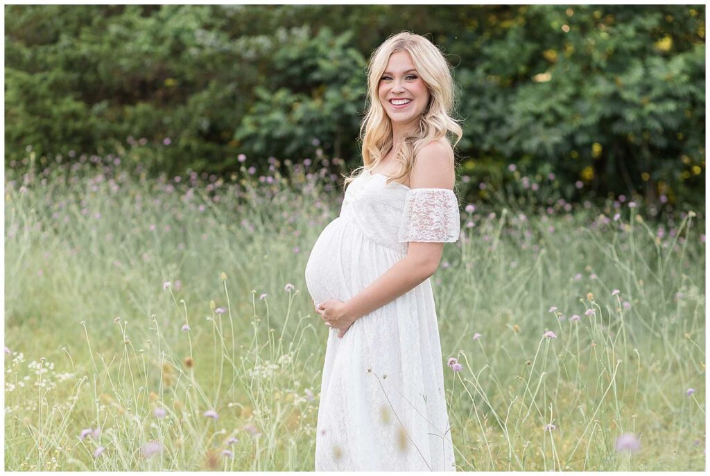 Maternity portrait captured by Wisp + Willow Photography has expecting mom-to-be standing in a field of purple wildflowers at Erwin Park and she stands sideways holding her belly and smiling at the camera.