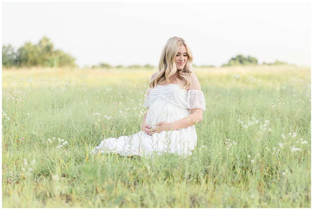 Gorgeous mom to be sits in a field of wildflowers at Erwin Lake in McKinney, TX wearing a white, off the shoulder, lace dress and holds her belly as she looks down off her shoulder.