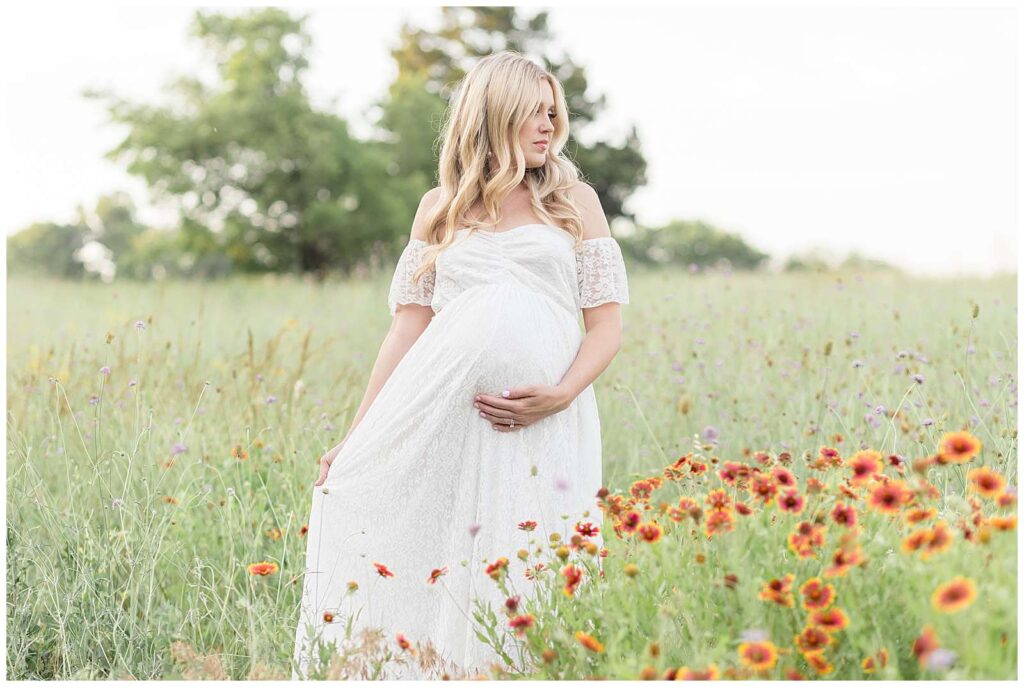Maternity portrait captures expecting mom standing in tall grass with blanket flowers as she looks off in the distance while holding her belly and wearing a white, off the shoulder, lace dress.