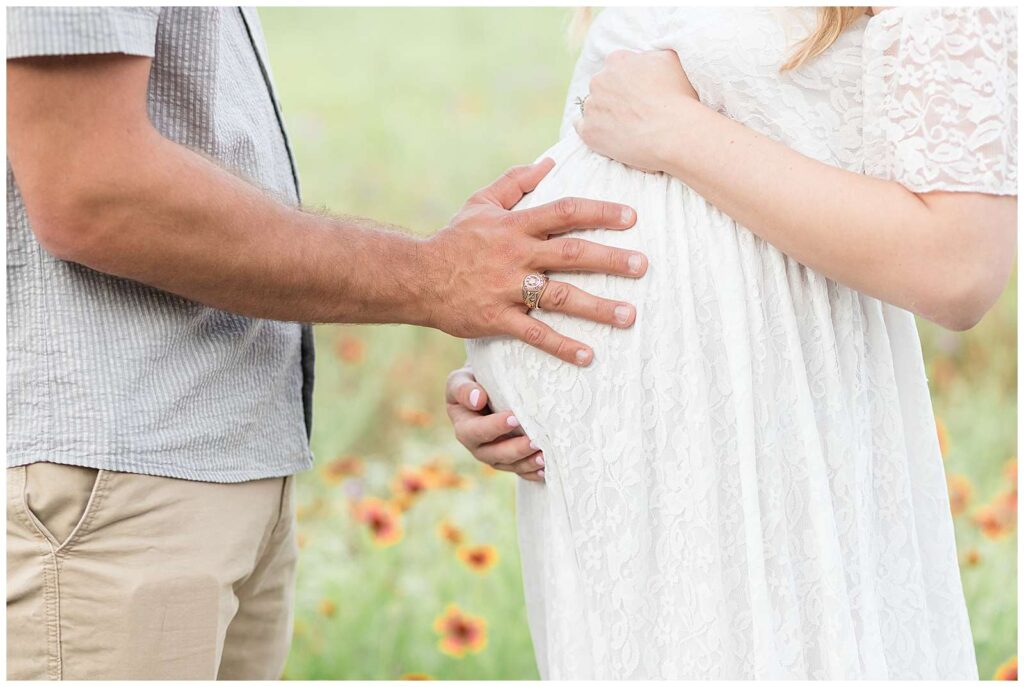 Close up image of a pregnant belly in a white lace dress,  has husband's hand holding her belly too with wildflowers in the background.