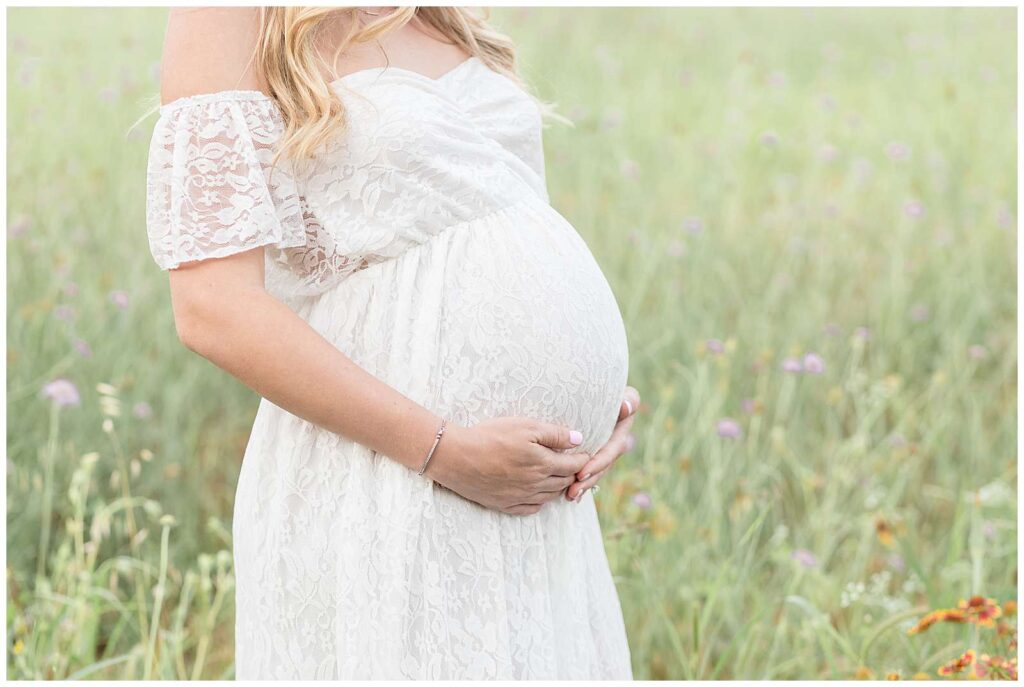 McKinney, TX maternity photographer takes beautiful images of a just a pregnant belly in a white, lace dress surrounded by a field of green and wildflowers.
