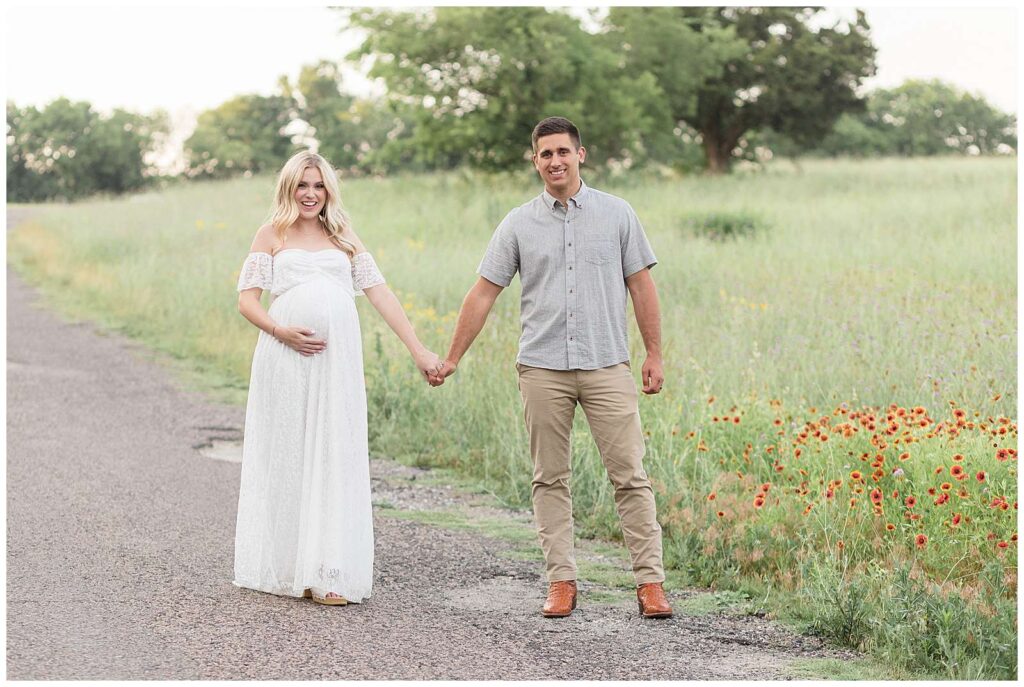 Expecting couple holds hands and walks down a path at Erwin Park in McKinney, TX with a field on one side filled with blanket flowers smiling at the camera of Wisp + Willow Photography Co.  See more of this session on the blog today!