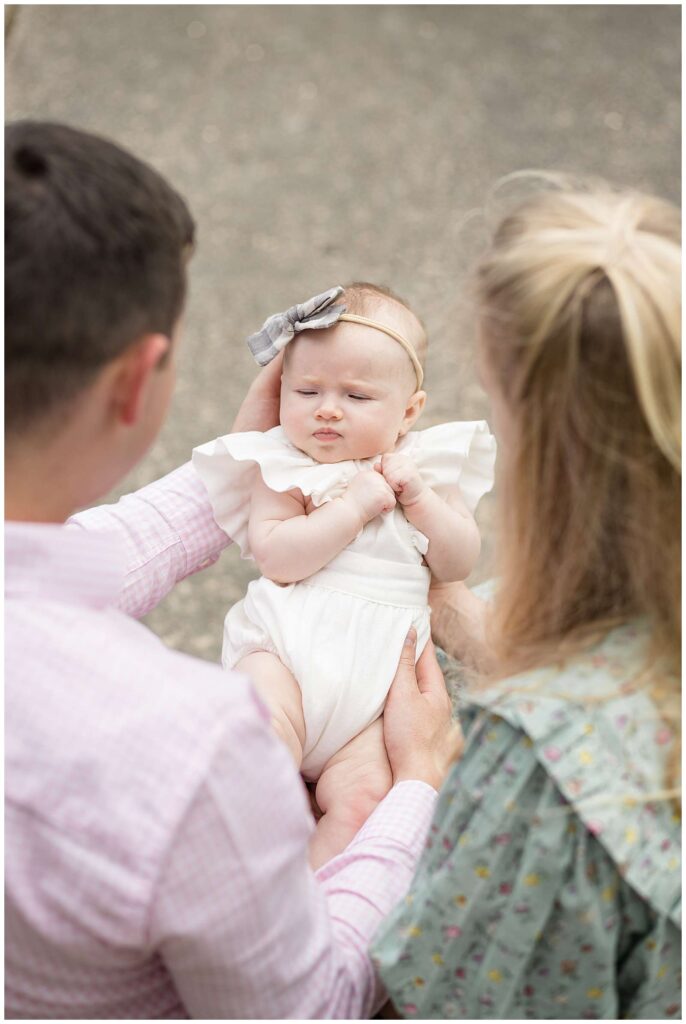 New parents hold their 3 month old daughter out in front of them as their family portrait photographer, Wisp + Willow Photography Co. stand above them and take the picture overhead focuses on their baby girl who wears a white onesie and headband.