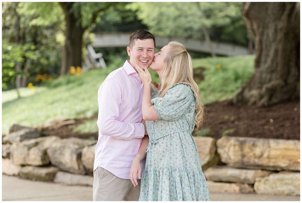 Young couple takes family portraits at Pullen Park in Raleigh, NC with Wisp + Willow Photography Co.  The Dad smiles at the camera as his wife pulls his head close to her and gives him a kiss on his cheek.