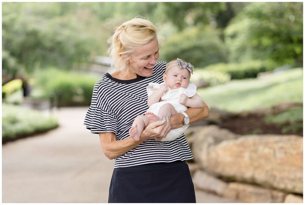 Proud Grandma holds her 3 month old granddaughter during a family portrait session at Pullen Park in Raleigh, NC.  She looks down at her granddaughter smiling happily!
