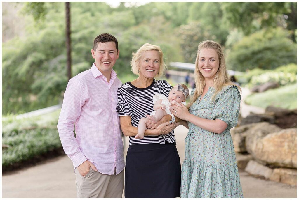 Raleigh family photographer, Wisp + Willow Photography Co., capture new parents, their 3 month old daughter, and her grandmother.  They stand in front of a walkway at Pullen Park with trees and a stone wall behind.