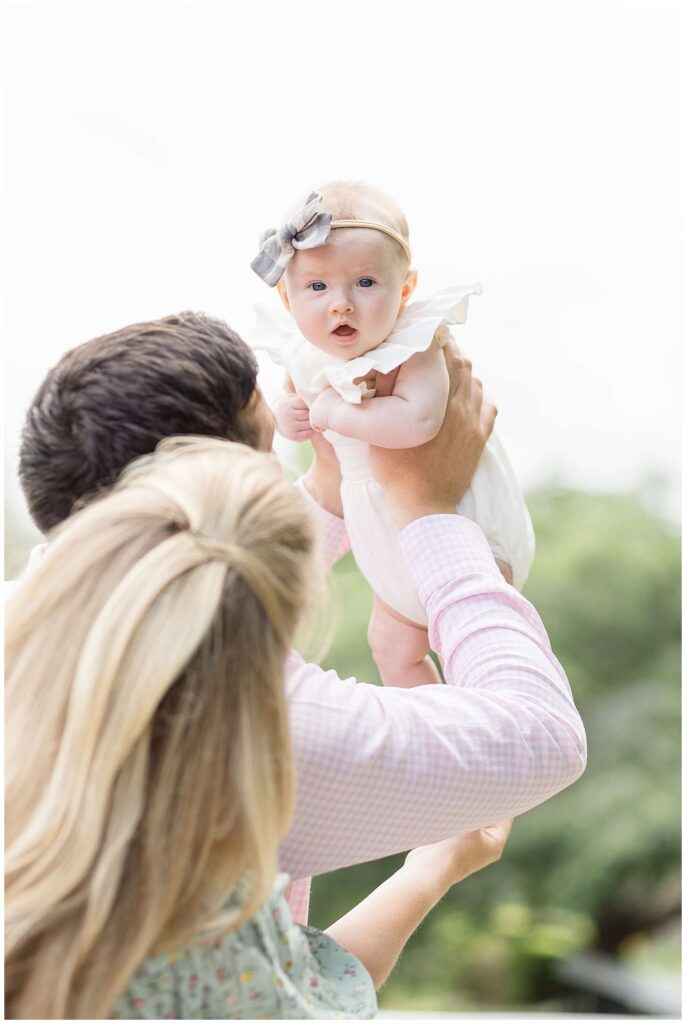New Dad lifts his 3 month old baby girl up into the air as she looks down to the camera of Wisp + Willow Photography Co.  She wears a white onesie and a headband bow for their family portrait session at Pullen Park.