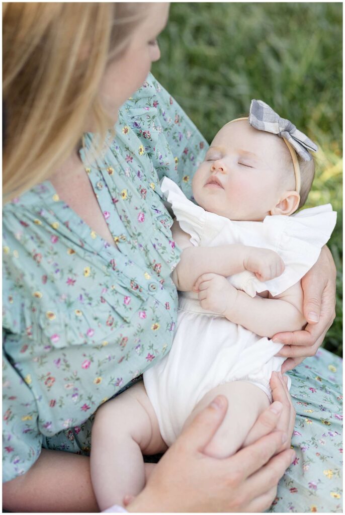 Young mom who is wearing a floral dress, holds her baby girl in her arms and looks down at her as she peacefully sleeps and wears a bow headband and white dress.