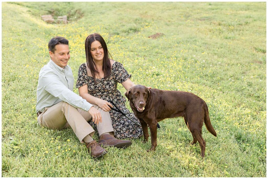 Couple sits in field with buttercups as the both look at their chocolate lab who stands slighting in front of them.  More from this Nashville portrait session on the blog today!