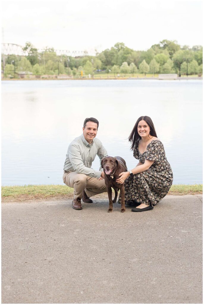 Couple bends down with their chocolate lab puppy in front of the lake at Shelby Bottom Nature Preserve during their photography session with Wisp + Willow Photography Co.