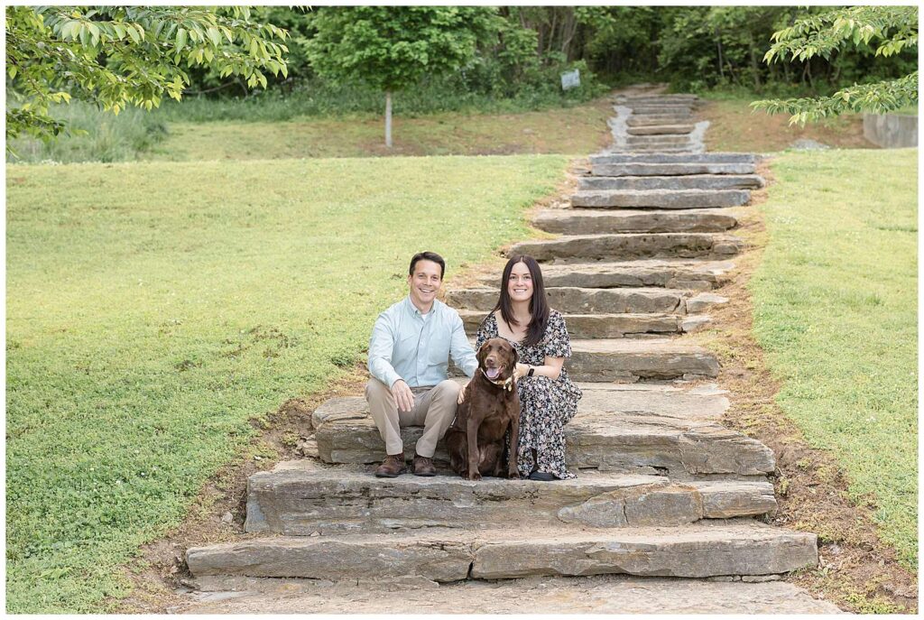Couple sits on a rock stair pathway down the fields at Shelby Botton Nature Preserve with their Chocolate Lab Dog and looks at the camera of Wisp + Willow Photography Co.!