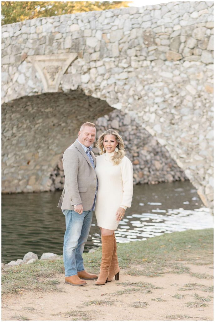 A Mom and Dad stand together during their fall family portraits at Adriatica Village as they smile at the camera wearing a cream colored sweater dress and suede boots while her husband wears jeans, a blazer, button down shirt, and boots.