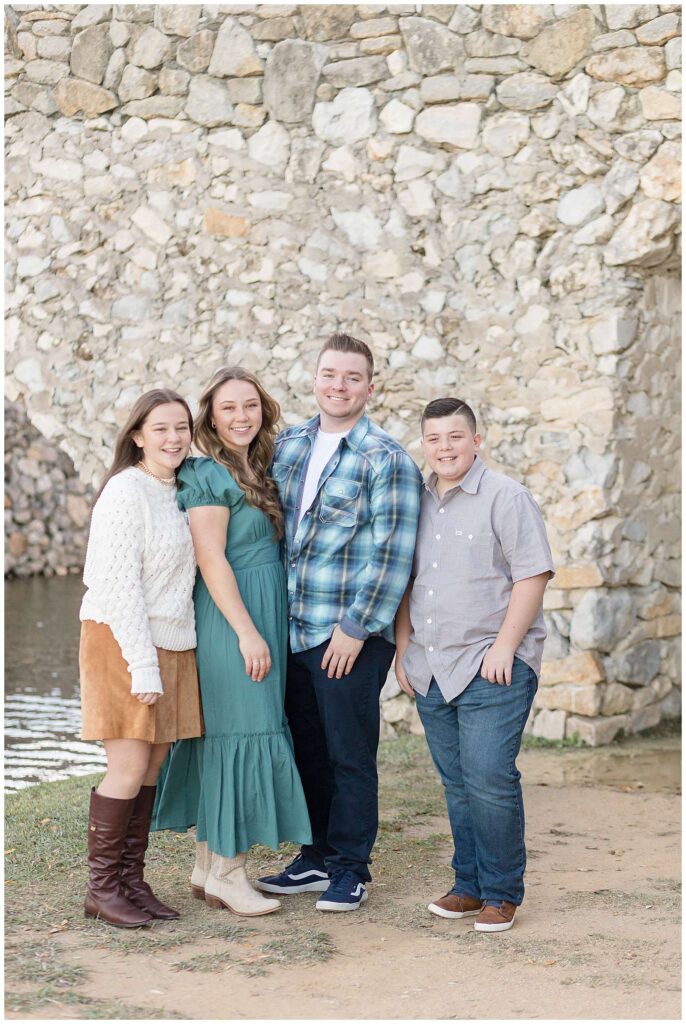 4 teenage siblings stand together in front of the stone wall of the Adriatica Village bridge in McKinney, TX for their fall family portraits.  