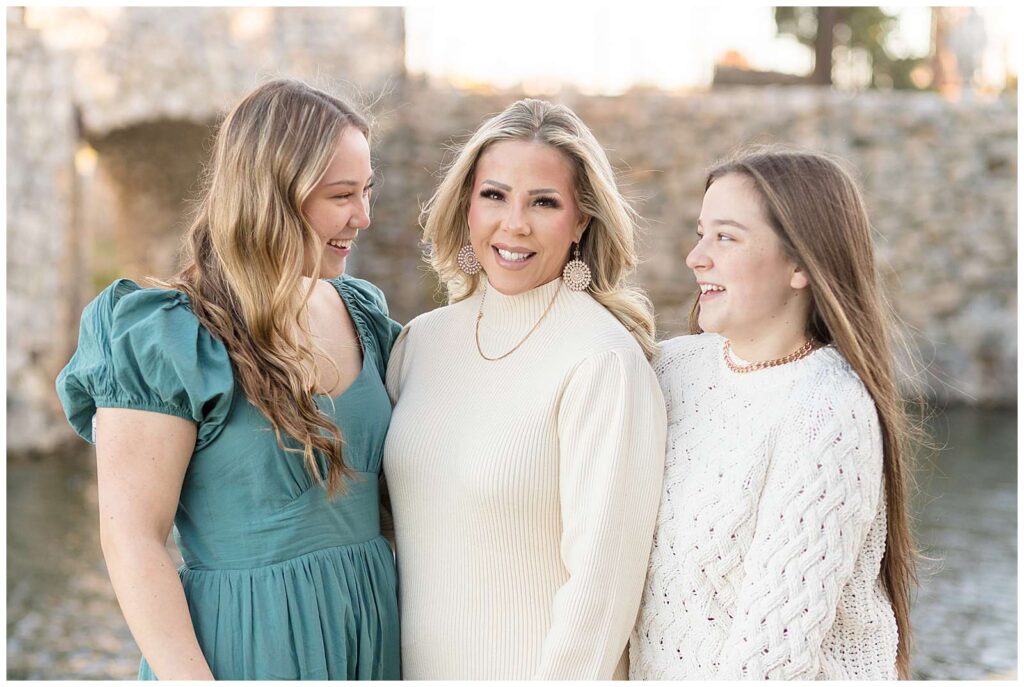 A mom, who is wearing a white, ribbed sweater stands in the middle of her teenage daughters who look at her and smile during their family portraits at Adriatica Village in McKinney, TX!