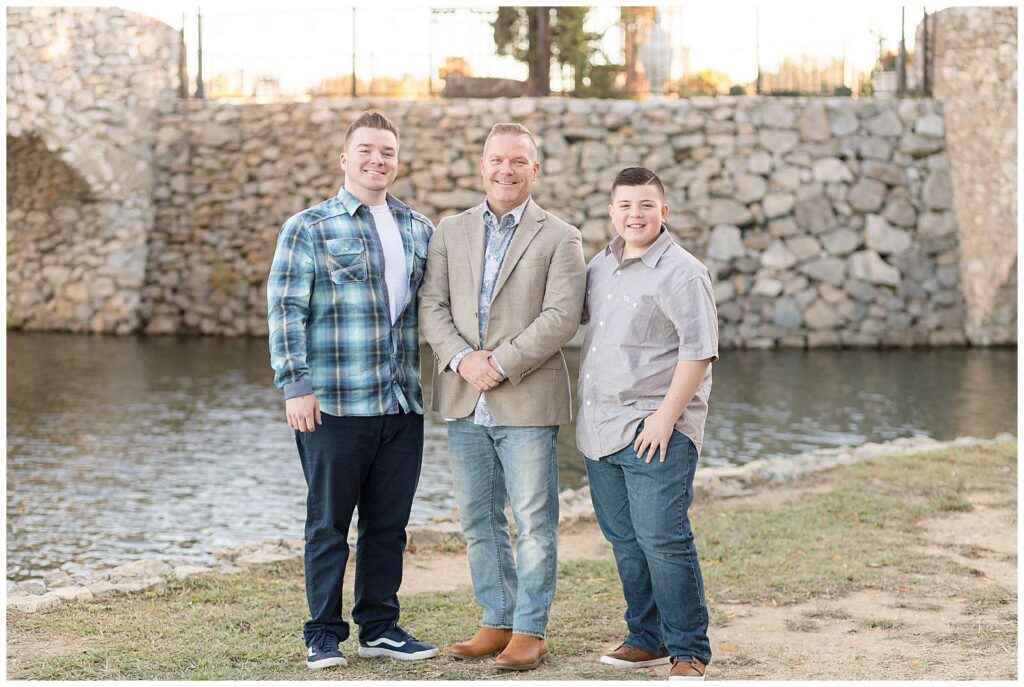 A Dad and his two sons stand together at Adriatica Village in front of the rock wall wearing coordinating colors of grey, blue, and khaki during their family session with Wisp + Willow Photography Co. 