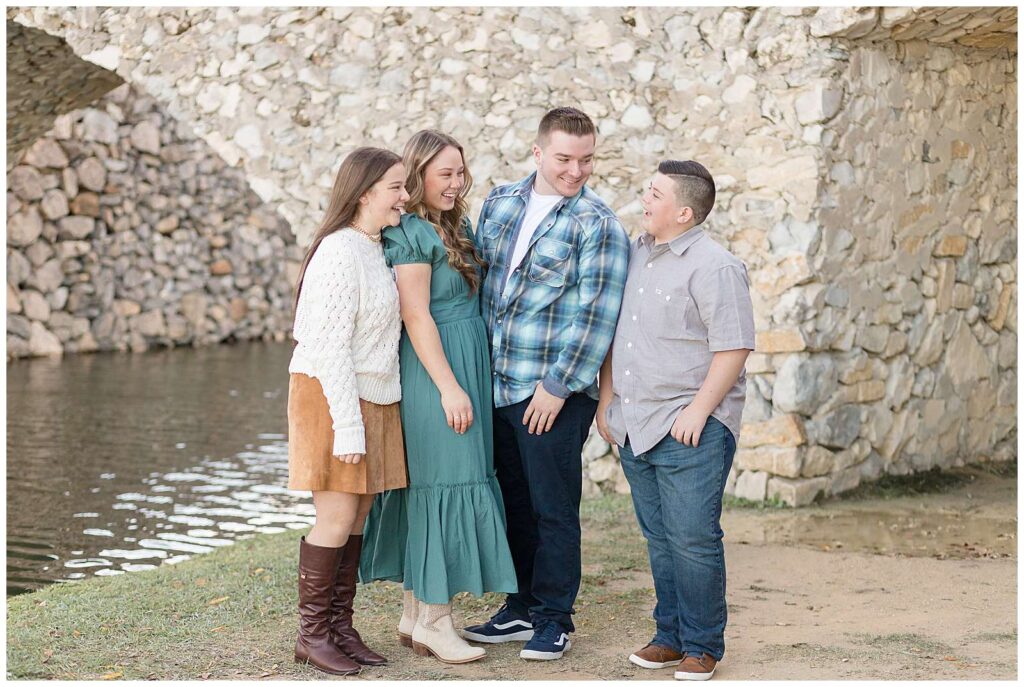 4 teenage siblings stand together in front of the Adriatica Village rock bridge and look at each other and smile and laugh for their fall family portraits!