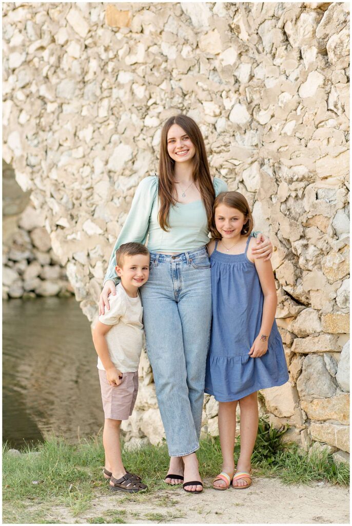 Adriatica Village family session has 3 siblings, 2 girls, and one boy, stand in front of the cobblestone bridge as the oldest stands in the middle and puts her arms around her brother and sister as they smile at the camera.
