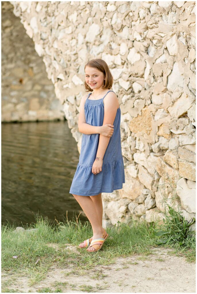 Young girl wears a light blue dress and rainbow sandals and stands in front of a rock bridge at Adriatica Village for a family session.