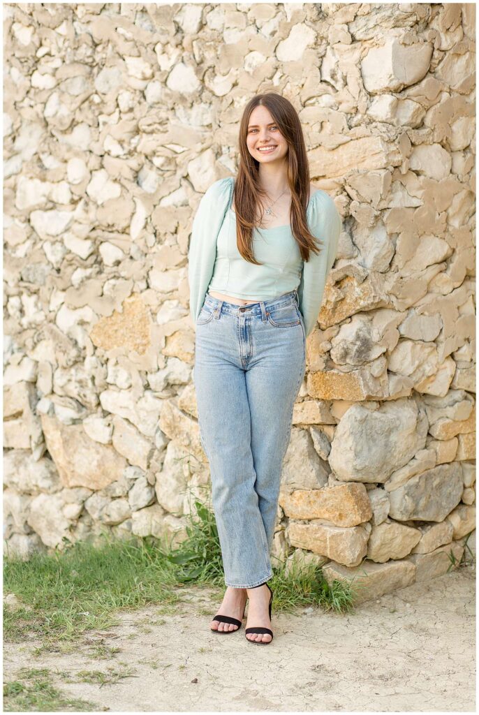 Teen girl wears jeans and a mint, long sleeve crop top and stands in front of a rock bridge at Adriatica Village and takes individual picture during their family session.