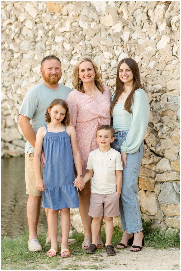 Family of 5 stand together in front of a rock wall at Adriatica Village as they take new family pictures.  They coordinate in light, pastel colors.