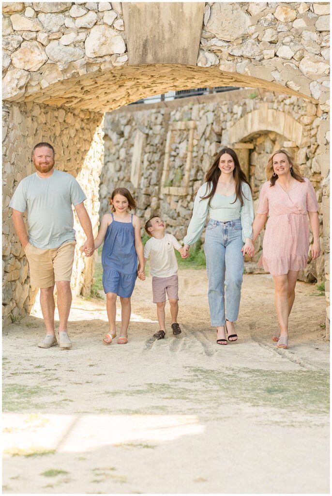 Family of 5 walk hand in hand and look at the camera smiling during their Adriatica Village Session with Wisp + Willow Photography Co.  Mom and Dad are on the ends with their kids in the middle.