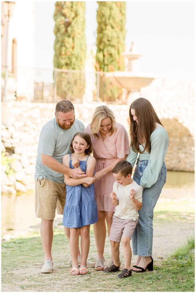 Family of 5 stand in front of the water and rock walls as they lean down and tickle their 3 kids.  They coordinate in light, pastel colors- light blue, mint, white, and pink.