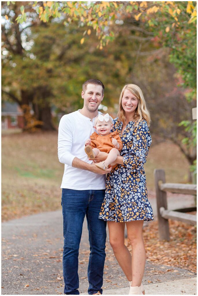 Family of 3 with toddler girl stand surround by fall leaves as Mom and Dad hold their daughter in between them during their family session in Raleigh, NC.