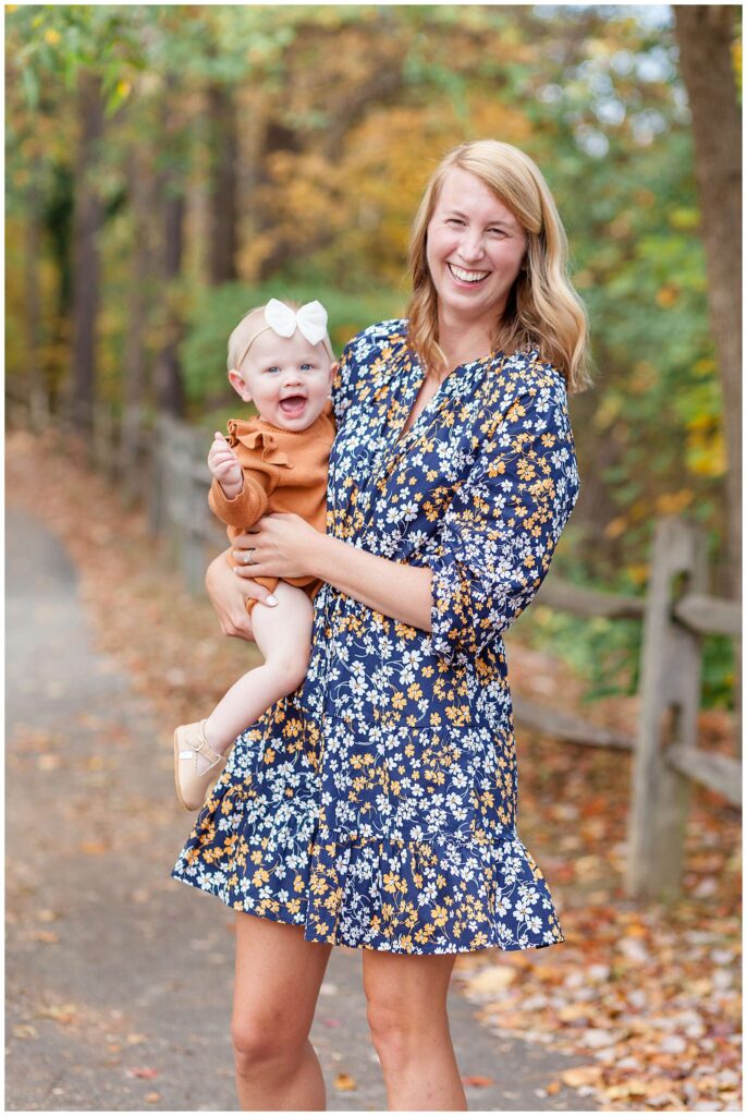 Mom and daughter take fall family portraits with Wisp + Willow Photography Co. in Raleigh, NC.  Mom wears a floral dress and her daughter coordinates with a rust, colored romper and a white bow headband.