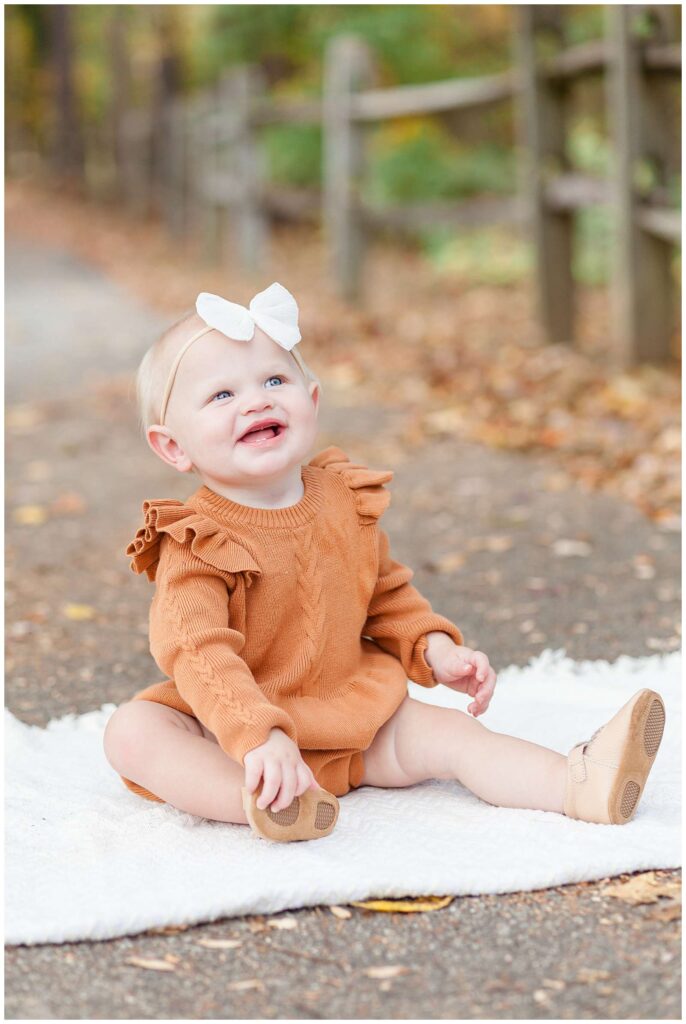 Baby girl sits on a white blanket wearing a rust, orange sweater romper and a white bow headband and smiles up to the right during family portraits in Raleigh, NC.