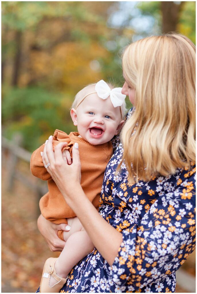 Mom holds her baby girl on her hip and looks down at her while her daughter smiles big with her two front teeth, white bow headband, and a burnt orange, sweater romper.