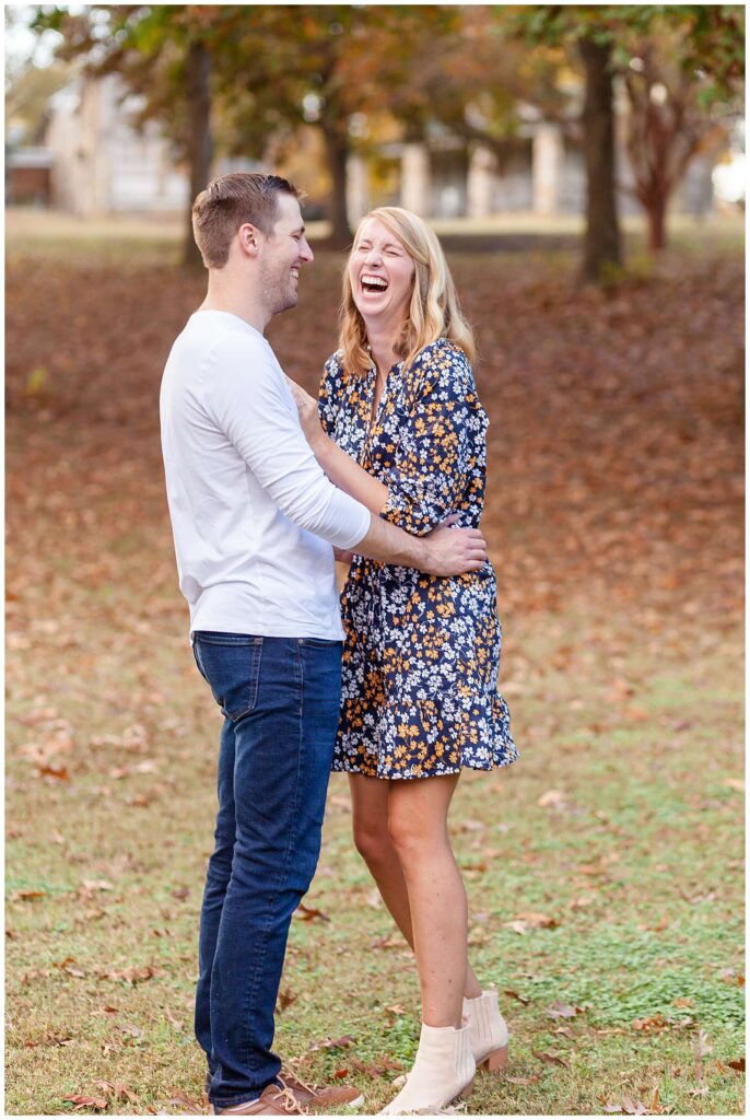 Couple stands in the grass surrounded with fall leaves on the ground as they come close to each other and the husband makes his wife laugh hysterically!  