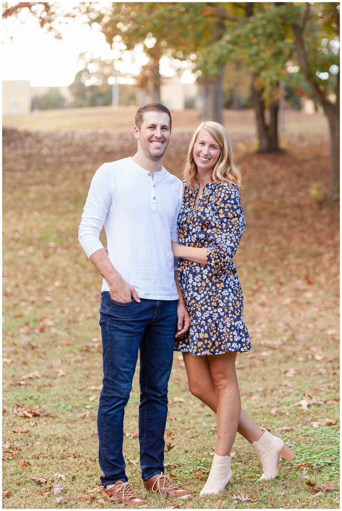 Couple stands in grass with fall leaves on the ground as they smile during their fall family photoshoot with Wisp + Willow Photography Co.  She wears a blue and orange floral dress and he wears a white henley shirt and blue jeans.