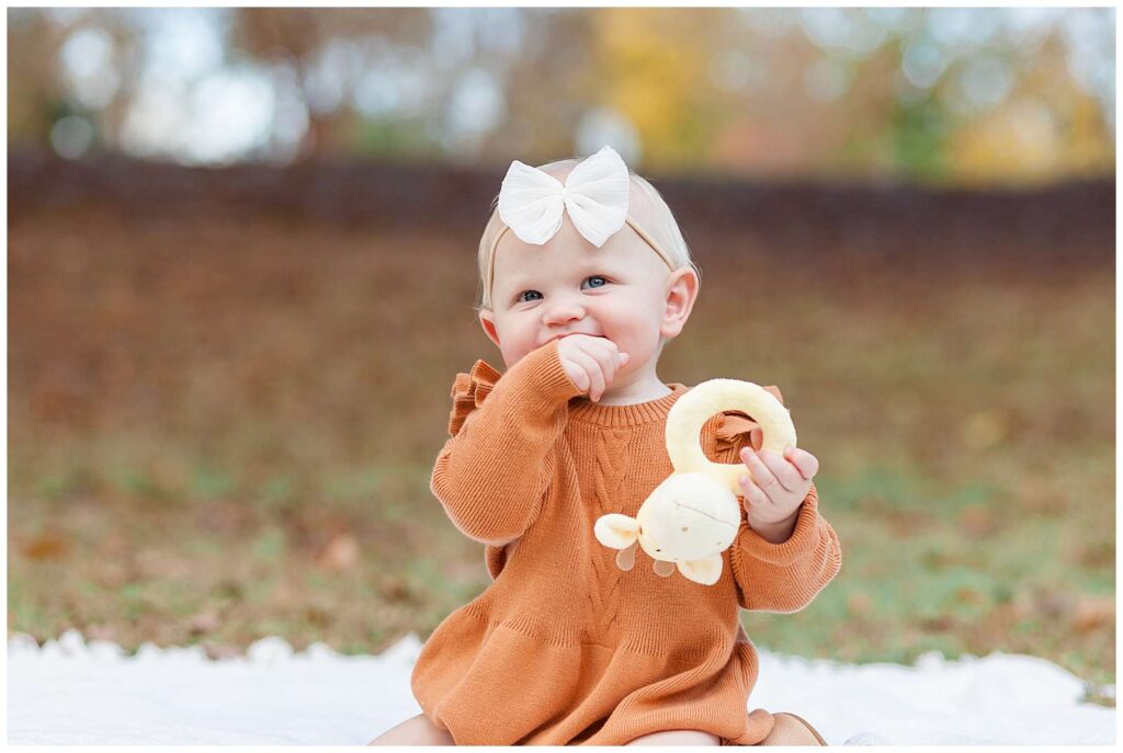 Baby girl sits on her knees on a white blanket with fallen fall leaves on the ground.  She wears a burnt, orange sweater romper, a white bow headband, and holds a soft, giraffe teether toy.