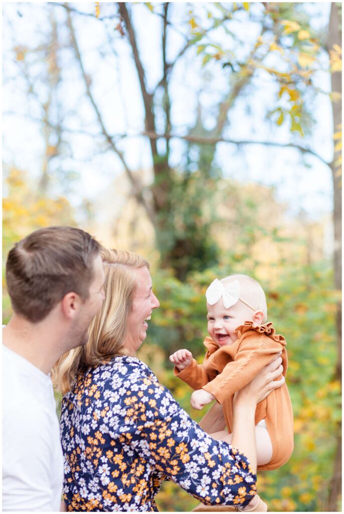 Wisp + Willow Photography Co. captures a family of 3 during their fall family session in Raleigh, NC.  Dad stands behind his wife as she holds their baby girl up in the air and she smiles back at them.  They coordinate in orange, blue, and white colored outfits.