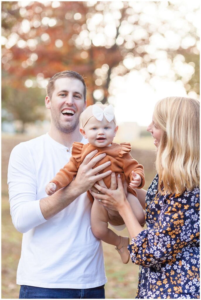 Mom and Dad hold their baby girl up in the air between them during their fall family photoshoot with Wisp + Willow Photography Co.  Mom is looking at her daughter while Dad and baby girl smile at the camera.  Click to see more of this family on the blog today!