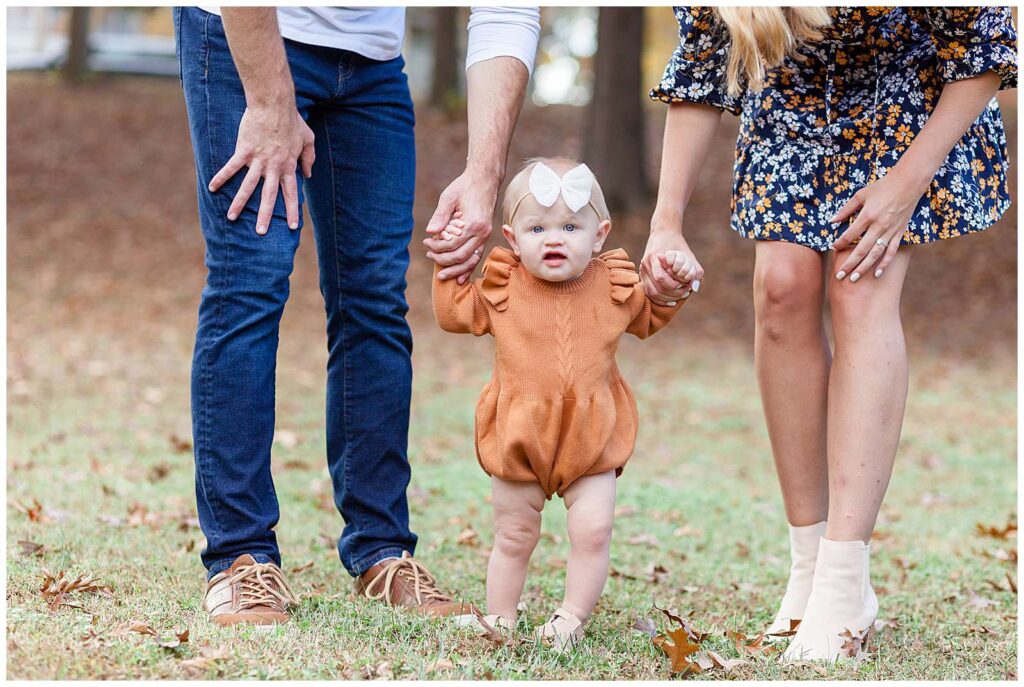 Mom and Dad hold their baby girls hands as she stands in between them and looks at the camera of Wisp + Willow Photography during their Raleigh fall family photography session.  The image is just of mom and dad's legs, feet and hands with the focus just on their baby girl who is wearing a burnt, orange sweater romper and a white bow, headband.