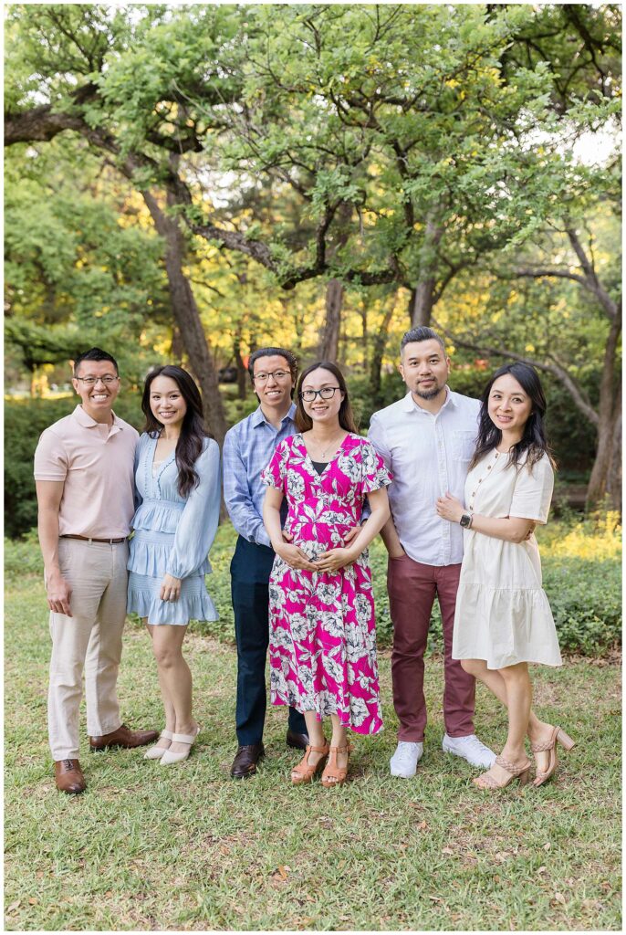 3 couples of an extended family portrait session at Davis Park in Dallas, TX stand together in front of trees with the glow of the sun behind them.