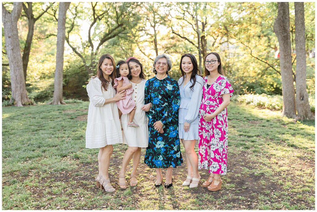 Grandma and the other 5 girls- 4 adults and one kid, stand together in front of the trees with the golden sun behind them for their extended family portrait session.  They all wear dresses with heels.