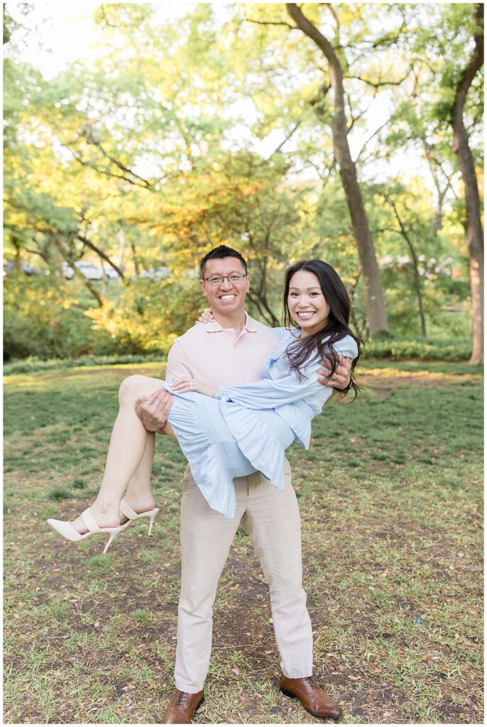 Couple stands on the grass in front of the trees with the golden sun coming through as he holds his wife in his arms and they both smile during their family portraits.  She wears a white, ruffle dress and cream heels and he wears a light pink polo shirt and khakis.