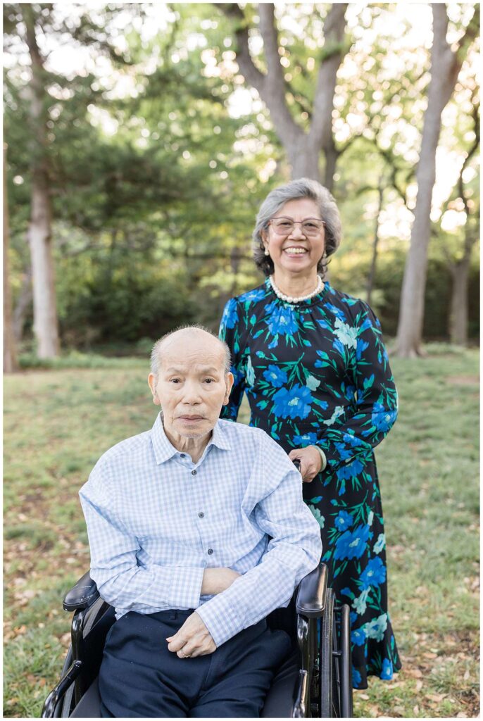 Elderly Mom and Dad coordinate wearing blue colors- Dad, who sits in a wheelchair wears a blue and white plaid shirt and Mom- who wears a blue and black floral dress, and a pearl necklace.