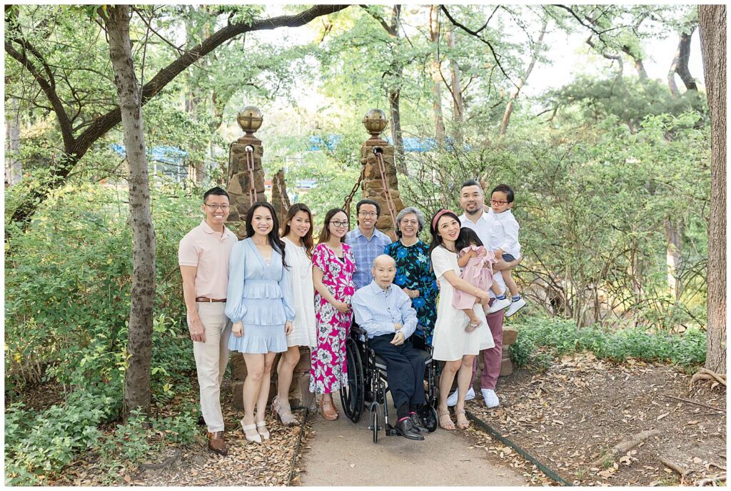 An extended family of 11 stand in front of a bridge at Davis Park in Dallas, TX coordinating in white, blue, and pink colors as they all stand behind their Grandpa/Dad in the wheelchair.