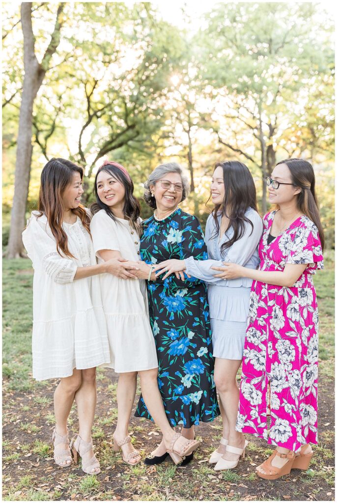4 daughters stand with their mom and they all look at each and laugh and hold each others arms during their extended family portraits at Davis Park in Dallas, TX.