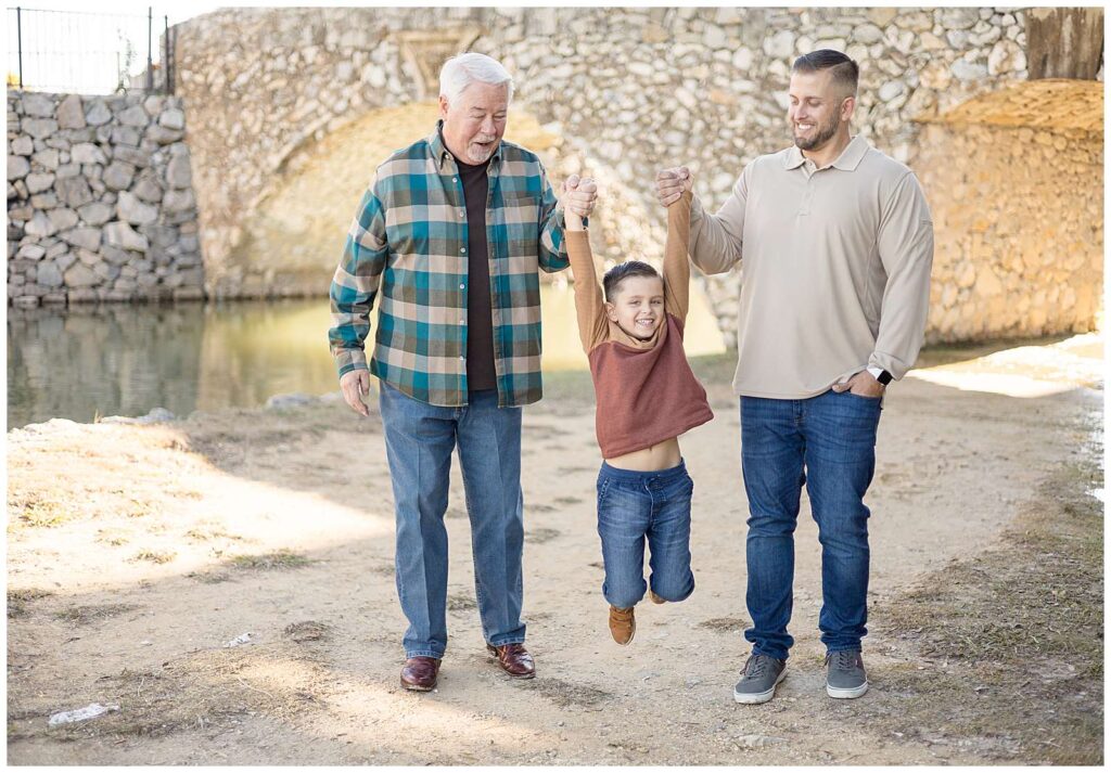 Grandpa, his grandson, and his dad swing their little boy in between them as they look down at him and the boy smiles at the camera during their extended family session at Adriatica Village.