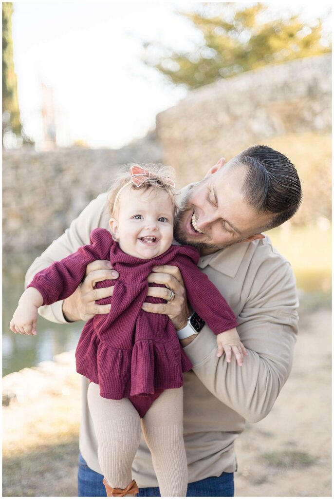A Dad, who wears a khaki shirt, holds his toddler daughter in front of him as he looks at her as she smiles big for the camera wearing a burgundy, fall dress and a peach, bow headband.