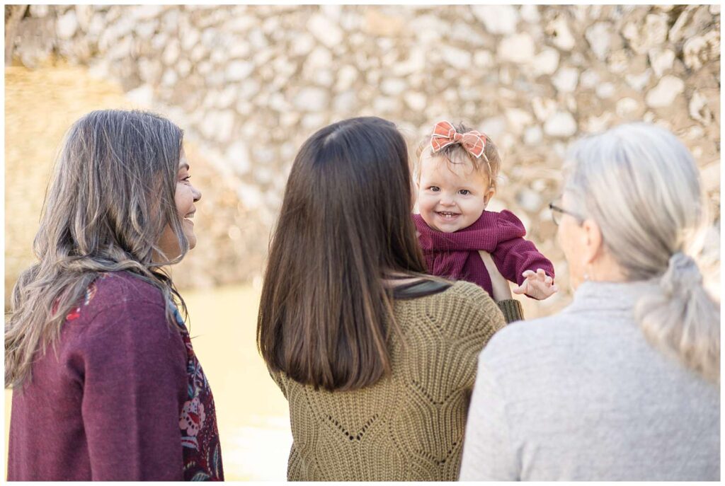 3 generations of women have their back to the camera as the youngest mom stands in the middle holding her baby girl up as she smiles at the camera.  