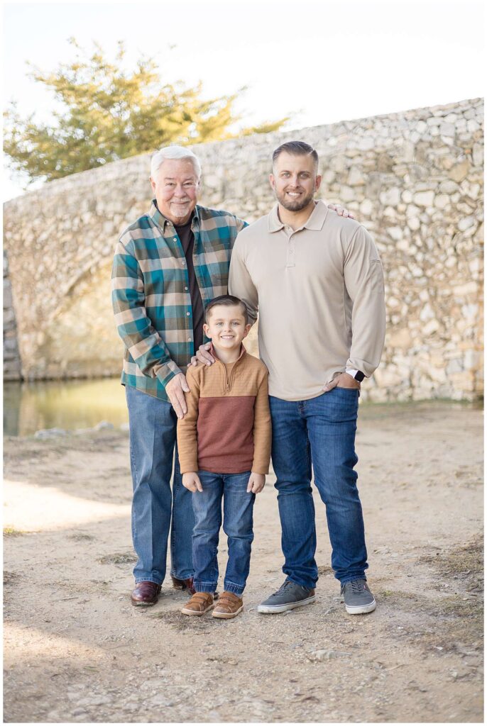3 generations of family men stand together wearing khaki, tan, and blue coordinating outfits and smile at the camera of Wisp + Willow Photography Co. 