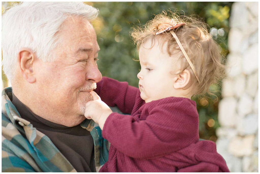 Grandpa holds his toddler granddaughter and they look towards each other as he smiles and she puts her finger on his lip.  A sweet grandfather and granddaughter moment.