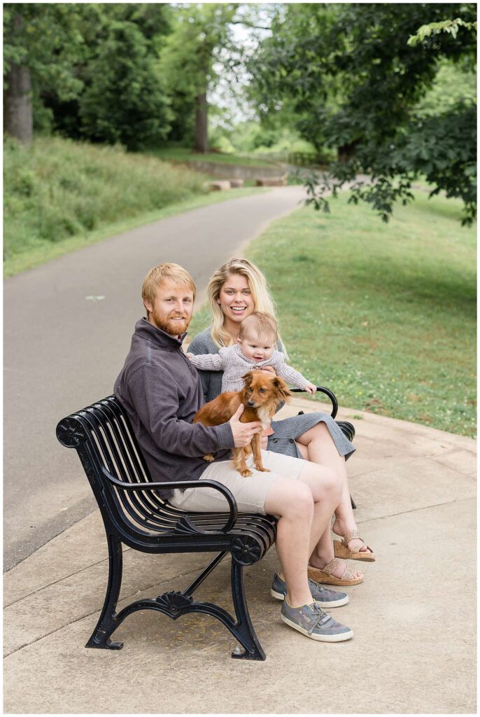 New parents sit on a park bench at Shelby Bottom Nature Preserve with their 6 month old standing in between them being held by mom and Dad holds their Dachsund dog on his lap.