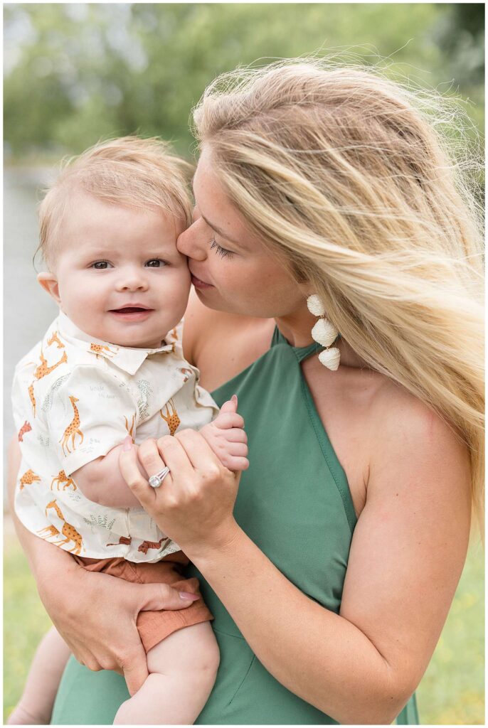 Mom who wears a green, sleeveless dress and white earrings, holds her 6 month old son who wears a shirt with giraffes all over, on her hip and holds his wrist and little baby hand as he smiles at the camera and she gives him a kiss on his cheek.